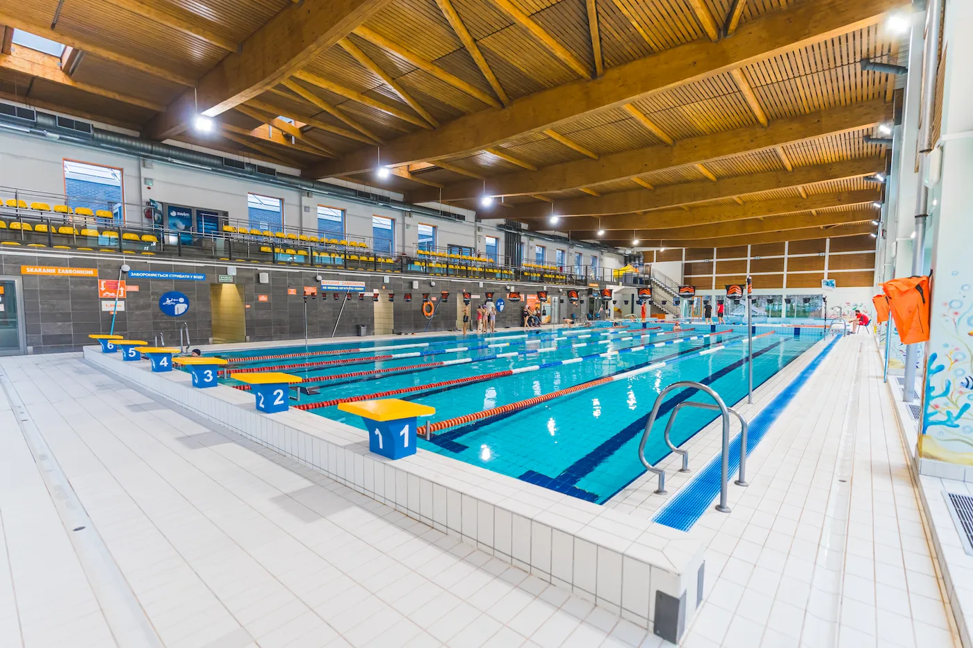 Indoor swimming facility with multiple competition swimming lanes, starting blocks numbered 1 through 6, spectator seating, tiled floors, and a large wooden roof structure.