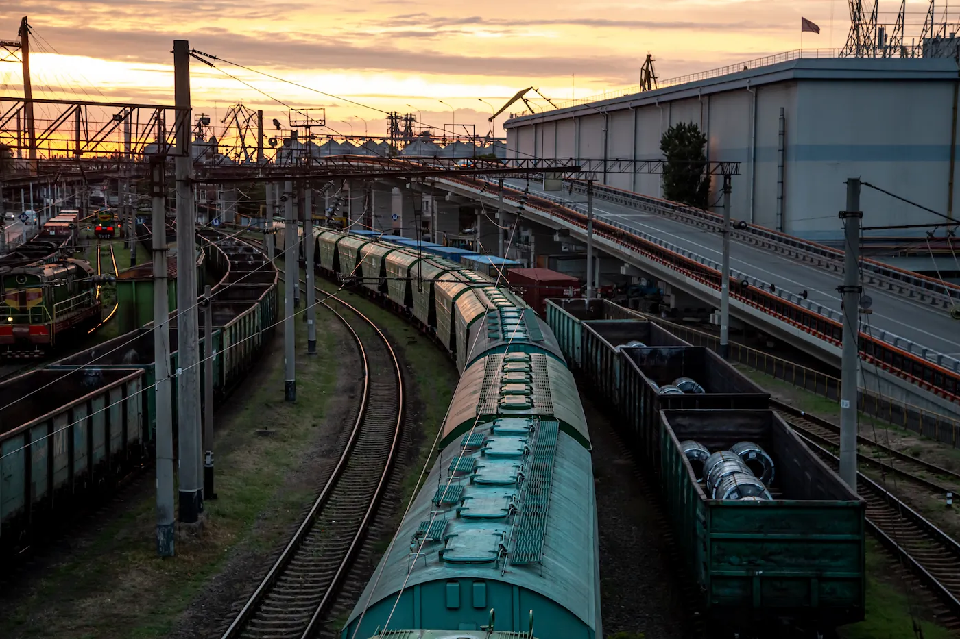 Rail freight yard with multiple cargo trains on parallel tracks, overhead electrical lines, freight wagons carrying industrial materials, and an elevated roadway beside the rail facility at sunset.