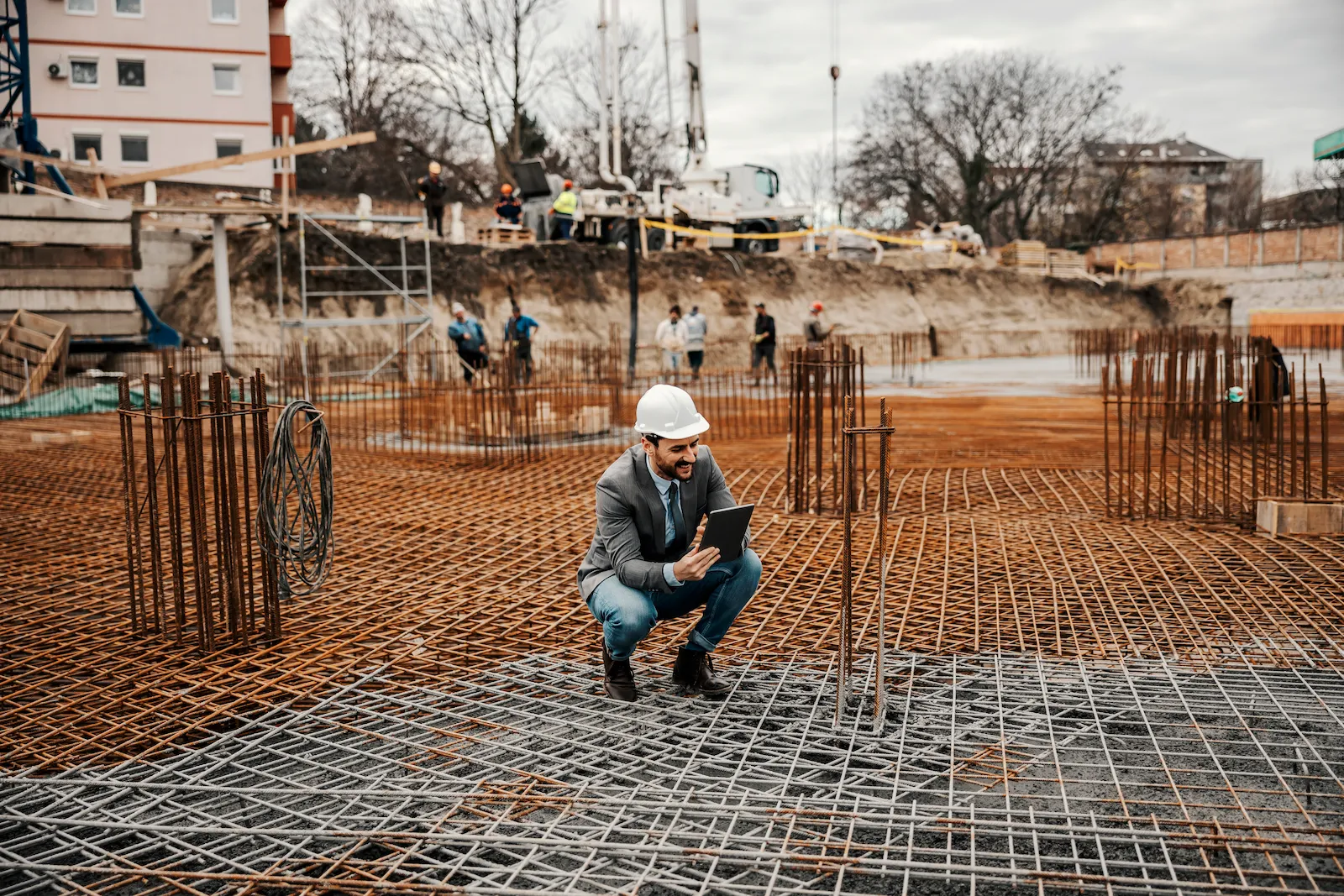 A civil engineer in a white hard hat and suit jacket squats on a steel rebar grid foundation while reviewing structural data on a digital tablet at an active construction site.