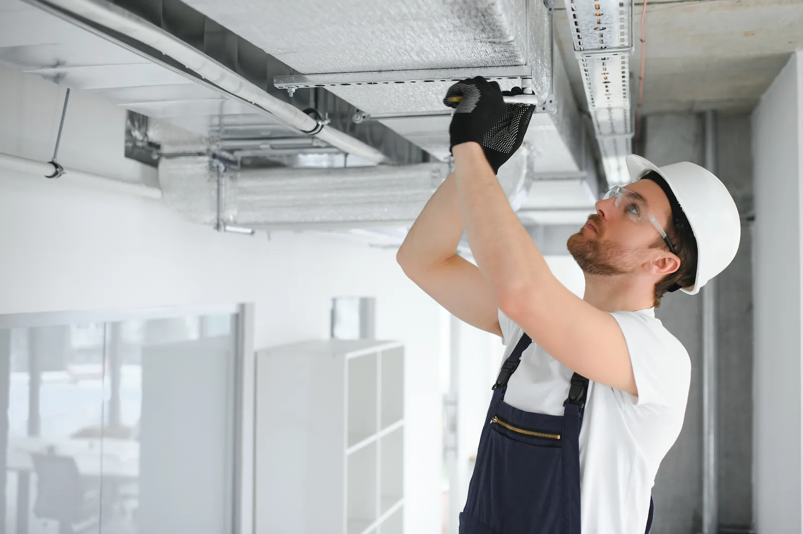 A professional HVAC engineer wearing a hard hat, safety glasses, and gloves inspects metal ventilation ductwork on an industrial ceiling.